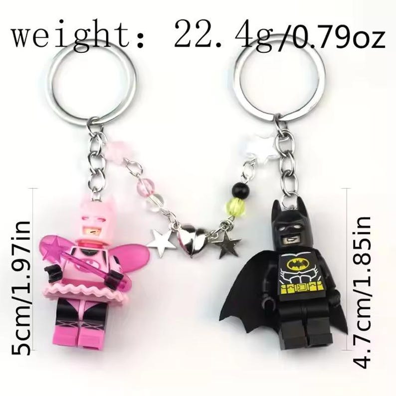May include: A pink and black Lego minifigure keychain with a silver chain and star charms. The pink minifigure is wearing a pink dress and wings. The black minifigure is Batman. The keychain is 5 inches long and weighs 22.4 grams.