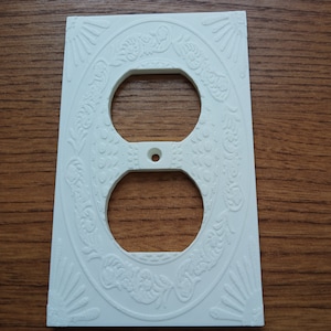 May include: White plastic outlet cover with an ornate embossed design. The cover has two standard outlet openings.