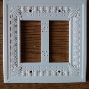 May include: White plastic light switch cover with an ornate design. The cover has two openings for switches and is designed to be mounted on a wall.