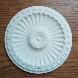 Może przedstawiać: Biały medalion sufitowy z ozdobnym, wypukłym, okrągłym wzorem. Środek medalionu zawiera mniejszy, wypukły, okrągły wzór z kwiatowym motywem.