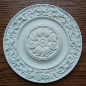Puede incluir: Medallón de techo blanco con un diseño floral. El medallón tiene un centro elevado con un diseño de flor y un borde de volutas intrincadas.