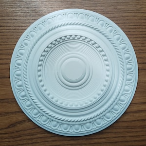 May include: A circular, pale blue ceiling rose featuring detailed, concentric patterns. The design incorporates rope, beaded, and floral motifs. The rose is made of a solid material and is set against a wood grain background.
