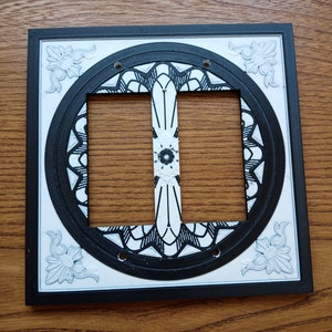 Puede incluir: Una tapa de interruptor de luz decorativa en blanco y negro con un diseño de mandala. La tapa es cuadrada con esquinas redondeadas y tiene un fondo blanco. El diseño de mandala está en el centro de la tapa y está rodeado por un borde negro.