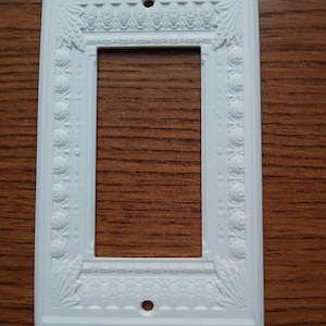 Könnte beinhalten: Weiße, dekorative Wandplatte aus Kunststoff mit einem verzierten Design. Die Platte hat eine rechteckige Öffnung für einen Lichtschalter.