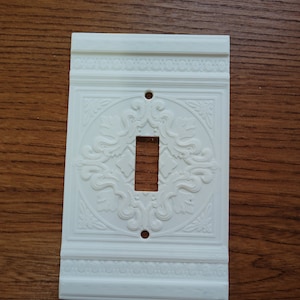 May include: White plastic light switch cover with an ornate, embossed design. The cover has a single switch opening.
