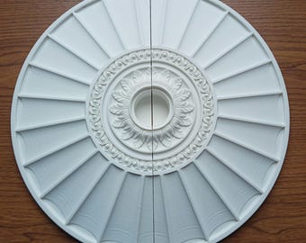 Könnte beinhalten: Eine weiße, kreisförmige Deckenrosette, geteilt in zwei Hälften, mit einem detaillierten floralen und geometrischen Design. Die Rosette hat ein zentrales Blumenmotiv, umgeben von Zierelementen und radialen Paneelen. Das Stück besteht aus einem matten, weißen Material.
