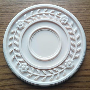 May include: A light pink, circular decorative element with a floral and leaf design. The design features a central circle, surrounded by a ring, a floral garland, and a raised edge. The piece is made of a solid, smooth material.