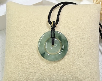 Può includere: Una collana con pendente in giada con una pietra verde circolare a forma di ciambella. La pietra è sospesa a un cordino nero. Il pendente poggia su un cuscino beige, con sfere decorative bianche e beige sullo sfondo.