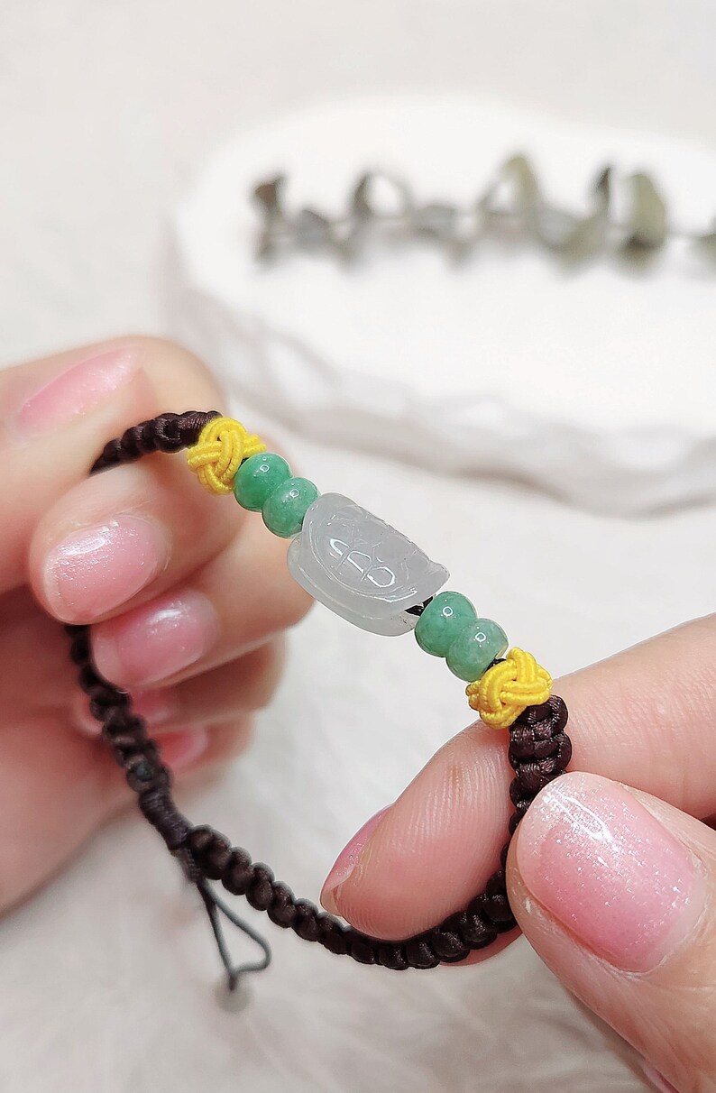 Peut inclure: Un bracelet en cordon marron avec des perles vertes et jaunes. Le bracelet est orn&eacute; d'un pendentif en jade avec un motif sculpt&eacute;.