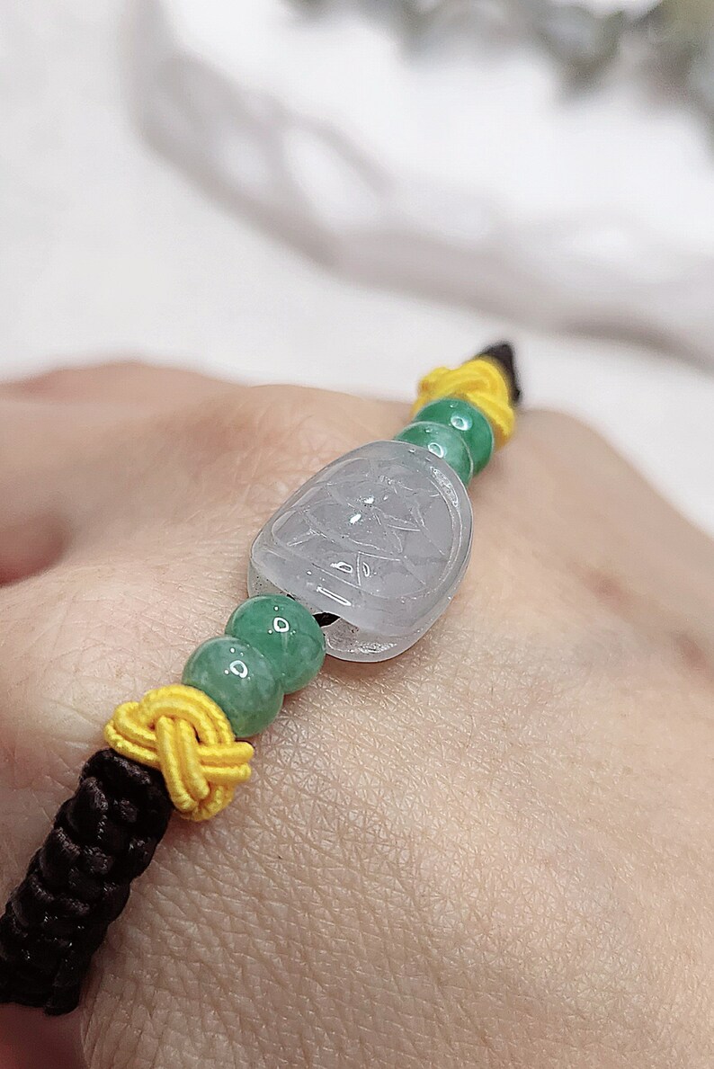 Peut inclure: Un bracelet en jade avec une perle de jade vert clair et un pendentif en jade blanc. Le bracelet est fait avec une ficelle brune et jaune.