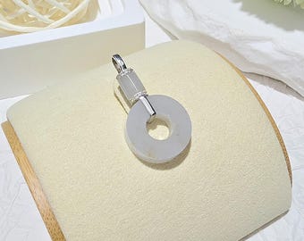 Può includere: Un pendente in argento con una pietra di giadaite circolare verde pallido. Il pendente ha un'ansa in argento e un piccolo accento rettangolare in giadaite. La giadaite ha un aspetto traslucido. Il pendente è esposto su una superficie color crema.