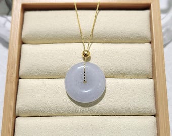 Può includere: Una collana con un ciondolo in giada lavanda chiaro, una catena d'oro e una perla dorata. Il ciondolo è rotondo e ha una superficie liscia. La collana è esposta in una scatola di legno con un rivestimento in velluto bianco.