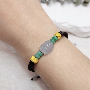 Peut inclure: Un bracelet en cordon noir avec des perles vertes et jaunes et un pendentif en jade gris clair. Le pendentif est orn&eacute; d'un motif sculpt&eacute;.