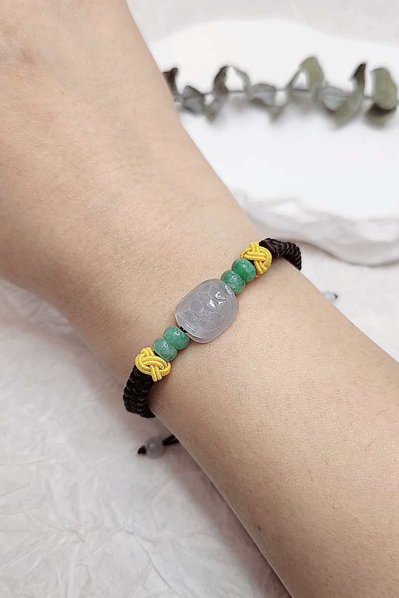 Peut inclure: Un bracelet en cordon noir avec des perles vertes et jaunes et un pendentif en jade gris clair. Le pendentif est orn&eacute; d'un motif sculpt&eacute;.