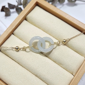 Pode incluir: Um delicado colar de corrente de ouro com dois anéis de jade entrelaçados. O colar é apresentado numa caixa de madeira com um forro de cor creme.