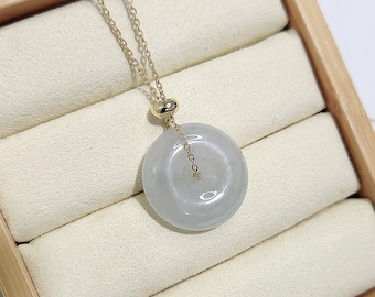 Puede incluir: Un delicado collar de cadena de oro con un colgante redondo de jade verde claro. El colgante es liso y pulido, y la cadena tiene un diseño simple y delicado.