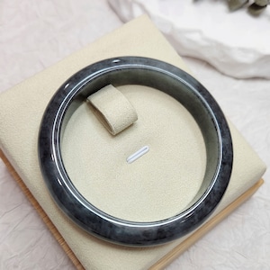 Peut inclure: Un bracelet en jade gris foncé sur un présentoir beige. Le bracelet est lisse et poli.