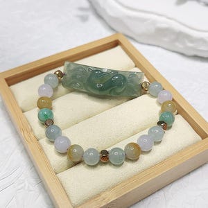 Puede incluir: Un brazalete de jade con un colgante de jade tallado en forma de dragón. El brazalete está hecho de cuentas redondas en tonos de verde, rosa y amarillo. El colgante es de color verde claro y tiene detalles intrincados.