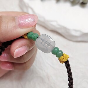 Peut inclure: Un bracelet en cordon tress&eacute; marron avec des perles vertes et jaunes et un charme en jade carr&eacute; avec des caract&egrave;res chinois.