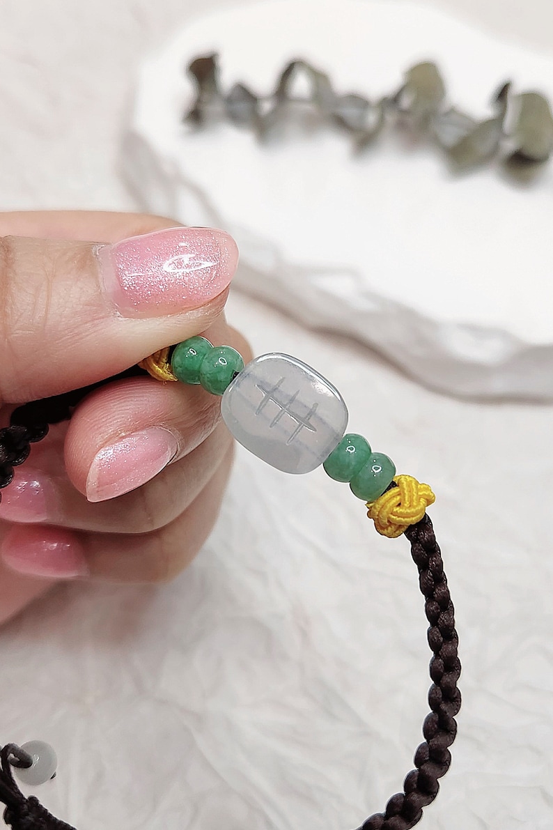 Peut inclure: Un bracelet en cordon tress&eacute; marron avec des perles vertes et jaunes et un charme en jade carr&eacute; avec des caract&egrave;res chinois.