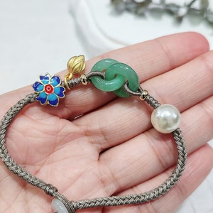 Puede incluir: Un brazalete de cordón trenzado marrón con un nudo de jade verde, un dije de flor de esmalte azul y rojo, un dije de hoja de oro y una perla blanca.