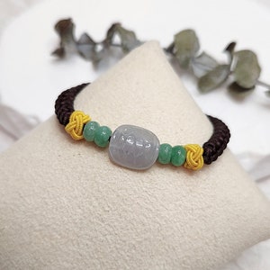 May include: A brown, yellow, and green braided bracelet with a light gray jade bead. The bead has a carved design.
