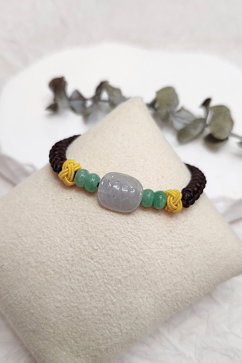 Peut inclure: Un bracelet tress&eacute; brun, jaune et vert avec une perle de jade gris clair. La perle est orn&eacute;e d'un motif sculpt&eacute;.
