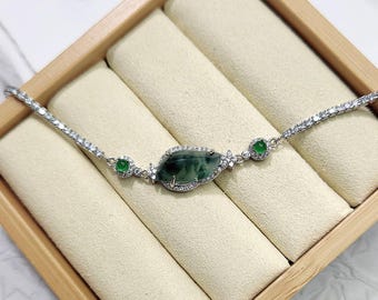 Op de afbeelding: Een zilveren armband met een centrale, donkergroene, ovaalvormige steen, omringd door kleinere, heldere stenen. Twee ronde groene stenen flankeren de centrale steen. De armband wordt gepresenteerd in een houten sieradendoos met een crèmekleurig interieur.