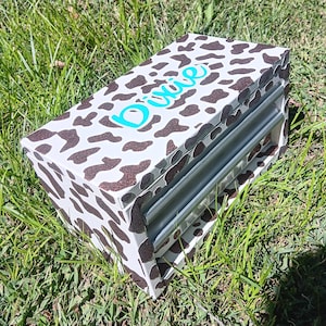 Puede incluir: Una caja de almacenamiento rectangular con un diseño de estampado de vaca en marrón y blanco. El nombre "Dixie" está escrito en turquesa en la parte superior. La caja tiene una ranura plateada en el lateral.