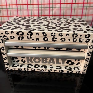 Puede incluir: Cofre de herramientas blanco con estampado de leopardo en blanco y negro. El cofre tiene dos cajones, con la palabra "KOBALT" impresa en gris en el cajón superior. Sobre una superficie negra.