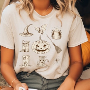 May include: A beige t-shirt with a Halloween theme featuring a pumpkin, witch's hat, bat, cauldron, broom, tombstone, tree, and spider web graphic.