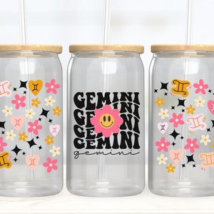 Puede incluir: Tres vasos de vidrio transparente con tapas de bambú y pajitas. Cada vaso presenta un diseño diferente. Uno tiene un patrón floral con corazones y estrellas, otro tiene la palabra "gemini" repetida en una fuente retro con una flor sonriente, y el último tiene un patrón floral con el símbolo del zodíaco Géminis.