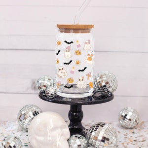 May include: A clear glass tumbler with a bamboo lid and straw, decorated with Halloween-themed illustrations of ghosts, bats, pumpkins, and flowers. The tumbler is displayed on a black cake stand with disco balls and a skull.