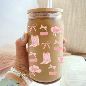 May include: A reusable glass tumbler with a brown lid and a straw. The tumbler is decorated with pink and white polka dot stickers of cherries, bows, cowboy boots, and hats.