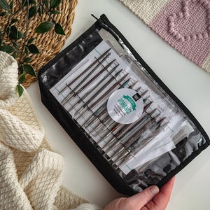 May include: A set of interchangeable knitting needles in a clear plastic case with a black zipper. The case has a label that says "Dyakcraft Pro Romance Interchangeable Knitting Needles".