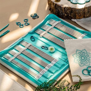 Op de afbeelding: Een set metalen breinaalden georganiseerd in een turquoise etui met rits. De naalden zijn gerangschikt in vakjes met labels die de maten 2mm US 0, 2.5mm US 1.5, 3mm US 2.5, 3.5mm US 4 en 5mm US 8 aangeven. Turquoise steekmarkeerders en een klein wit bloemetje zijn inbegrepen.