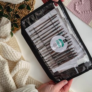 May include: A black zippered case with a clear plastic window containing a set of interchangeable circular knitting needles. The needles are labeled with sizes in millimeters. The case has a green and white label that reads "DROPS PRO ROMANCE Interchangeable Circular Needles Deluxe Set".