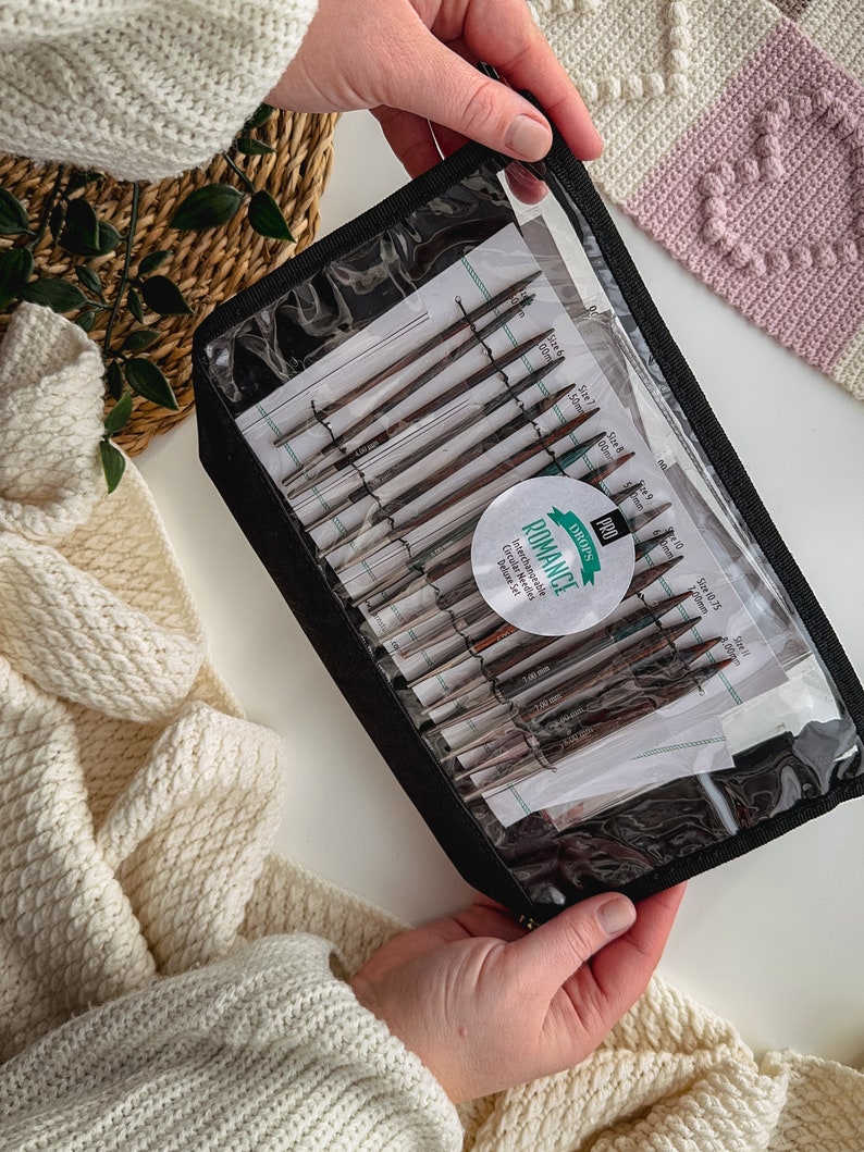 May include: A black zippered case with a clear plastic window containing a set of interchangeable circular knitting needles. The needles are labeled with sizes in millimeters. The case has a green and white label that reads "DROPS PRO ROMANCE Interchangeable Circular Needles Deluxe Set".