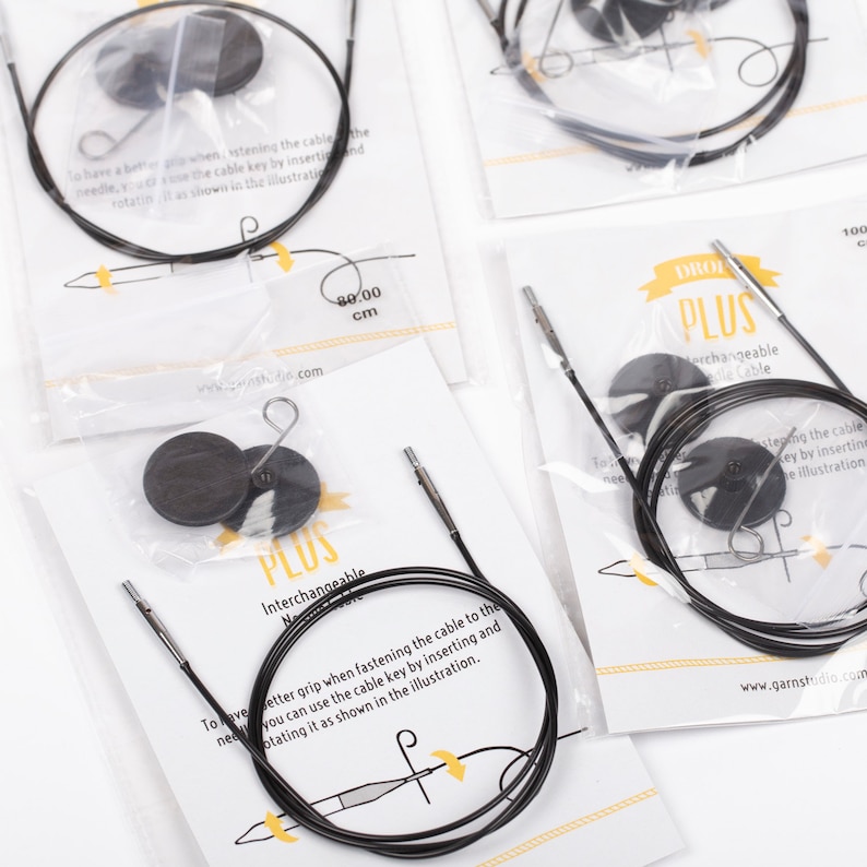 May include: Close-up of a set of black interchangeable knitting needles with a cable key. The needles are packaged in clear plastic bags with white labels that say "DROL PLUS Interchangeable Needle Cable".