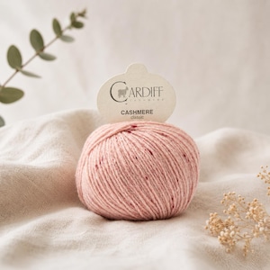 Könnte beinhalten: Ein Knäuel rosa Cardiff Cashmere Classic Garn mit gesprenkelter Textur. Das Garn wird auf einem cremefarbenen Stoff präsentiert, mit einem Etikett mit der Aufschrift "Cardiff Cashmere" und "Classic".