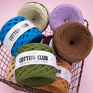 Peut inclure: Un panier en fil de fer rempli de pelotes de fil "Cotton Club Premium T-Shirt Yarn" colorées, dans des tons de bleu, vert, marron, violet et beige. Le fil est disposé en forme circulaire, avec les étiquettes visibles sur certaines pelotes. Le fond est rose clair.