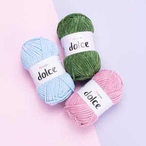 Peut inclure: Trois pelotes de fil doux dans des tons pastel : bleu clair, vert et rose. Chaque pelote porte une étiquette blanche avec le mot "dolce" en noir. Le fil est présenté sur un fond rose et violet.