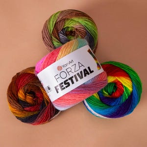 Può includere: Tre gomitoli di filato colorati sono disposti su una superficie marrone chiaro. Un gomitolo è etichettato "FORZA FESTIVAL" e presenta un arcobaleno di colori. Gli altri due gomitoli mostrano un mix di colori, tra cui verde, marrone, rosso e blu. Il logo YarnArt è visibile.