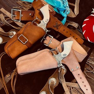 May include: An assortment of cowboy-themed items, including leather holsters, belts, and toy revolvers. The holsters are various shades of brown, with silver buckles and decorative accents. A turquoise bandana is visible in the background, adding a pop of color to the rustic theme.