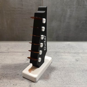 Może przedstawiać: Czarny stojak na kostki gitarowe z białą podstawą. Stojak ma kształt gitary, a na nim nadrukowane jest słowo "Gitara". W stojaku znajduje się kilka brązowych kostek gitarowych.