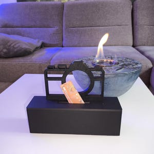 Pode incluir: Um cofre preto em forma de câmera com uma ranhura para inserir notas. Uma nota é parcialmente visível na ranhura. Uma pequena lareira decorativa redonda azul-acinzentada com uma chama está atrás do cofre. Os objetos estão sobre uma mesa branca.