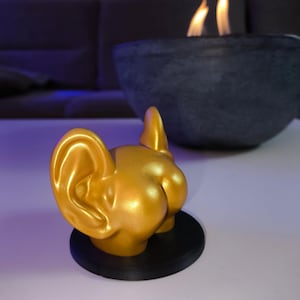 Könnte beinhalten: Eine goldfarbene Skulptur des Hinterteils eines Hundes, einschließlich der Ohren, ist auf einem schwarzen kreisförmigen Sockel montiert. Die Skulptur befindet sich vor einer dunkelgrauen Schale mit Flammen.