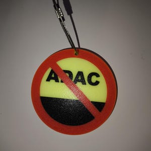 Op de afbeelding: Een ronde tag met een rode rand, geel en zwart midden, en het woord "ADAC" rood doorgestreept. Het is bevestigd aan een metalen kabel met een lus.