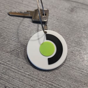 Op de afbeelding: Ronde witte sleutelhanger met een zwart en groen logo. Het logo is een cirkel met een zwarte halvemaanvorm en een groene cirkel erin.