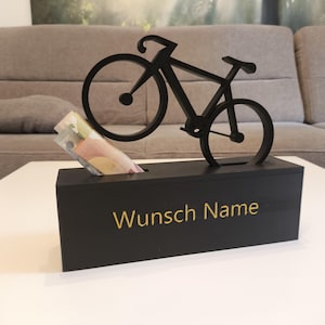 Könnte beinhalten: Schwarze Fahrrad-Spardose mit dem goldenen Schriftzug "Wunsch Name". Die Spardose hat einen Schlitz zum Einwerfen von Geldscheinen, und einige Scheine sind sichtbar. Das Fahrrad ist oben auf der Box montiert.